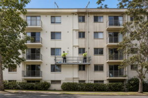 Painters on a suspended platform repainting the rendered facade of a Sydney apartment building, with the finished and unfinished sections both visible